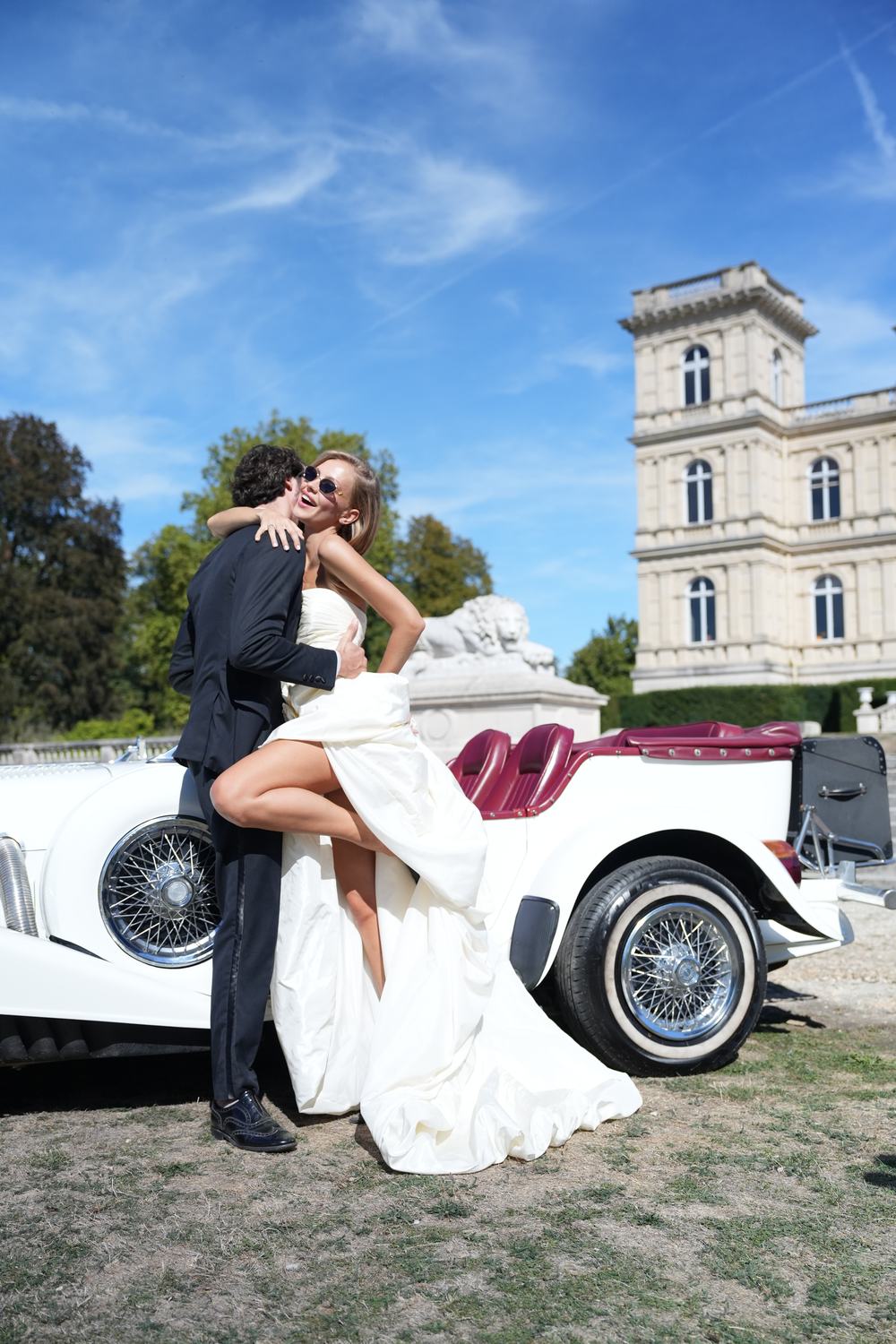 La mariée, rieuse, saute dans les bras du marié devant la voiture ancienne et la tour du château