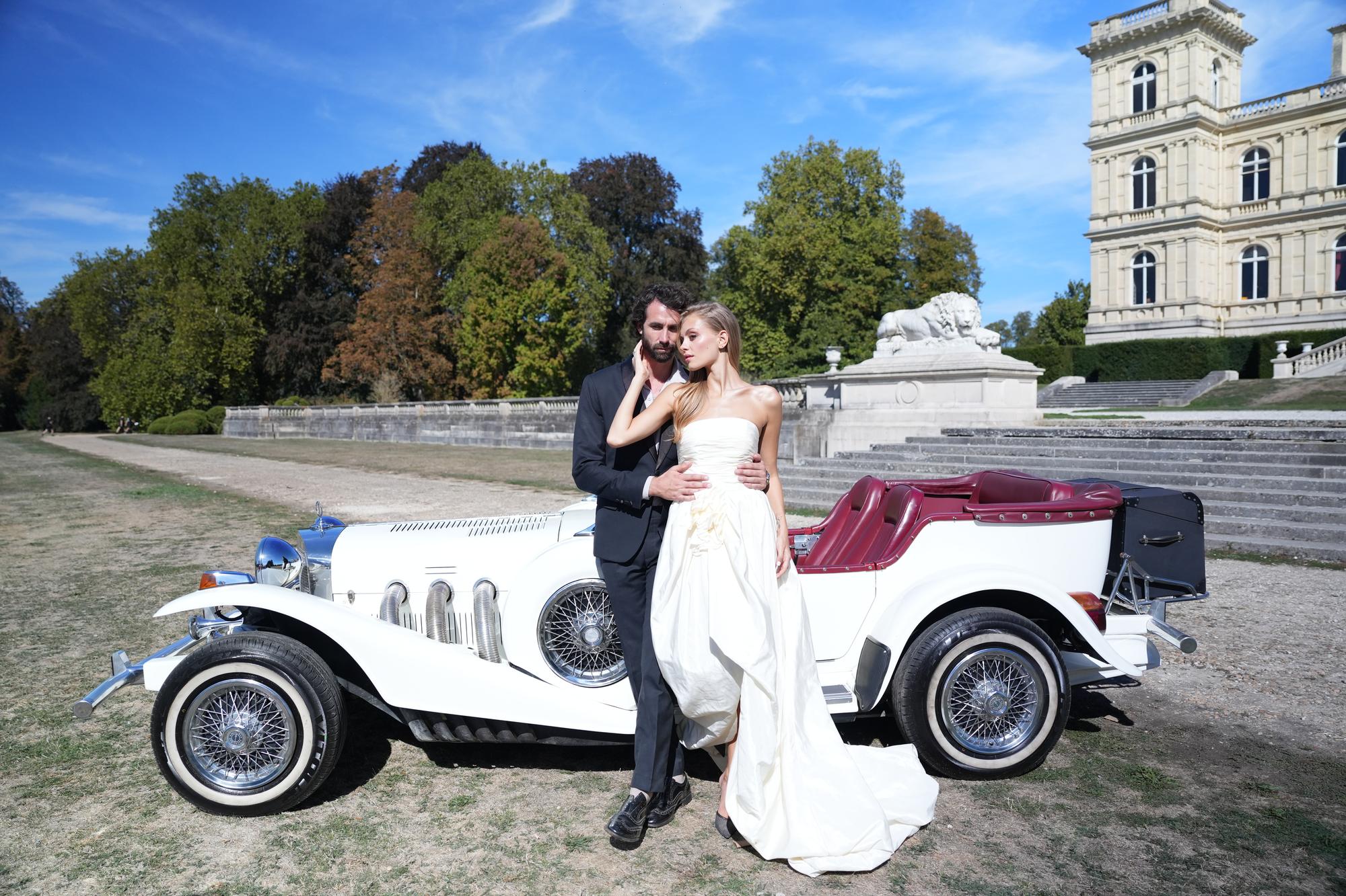 Vidéaste de mariage : les mariés enlacés devant une voiture ancienne blanche, dans le parc d'un château